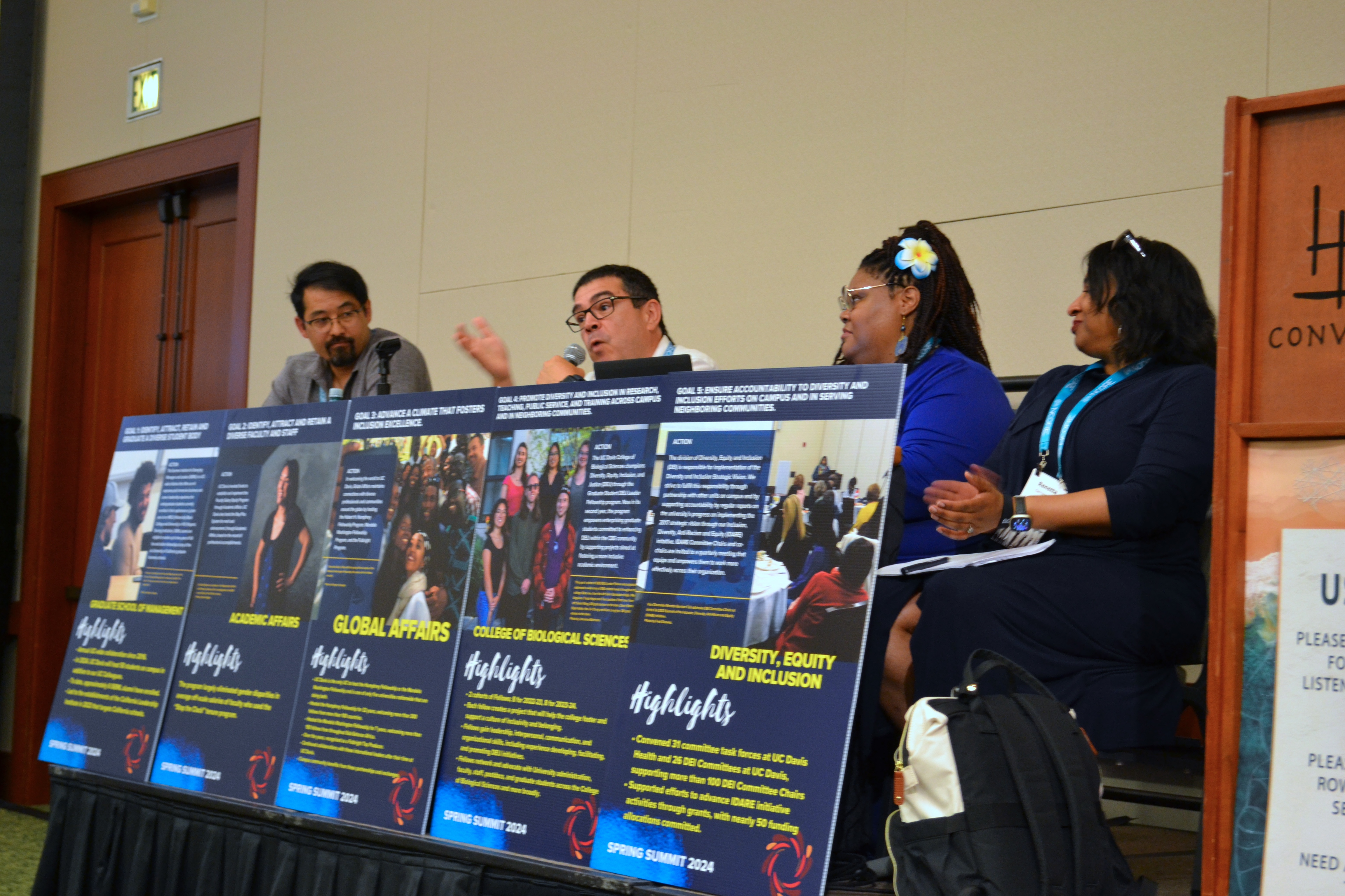 Four panelists seated behind a table with five posters at a conference.