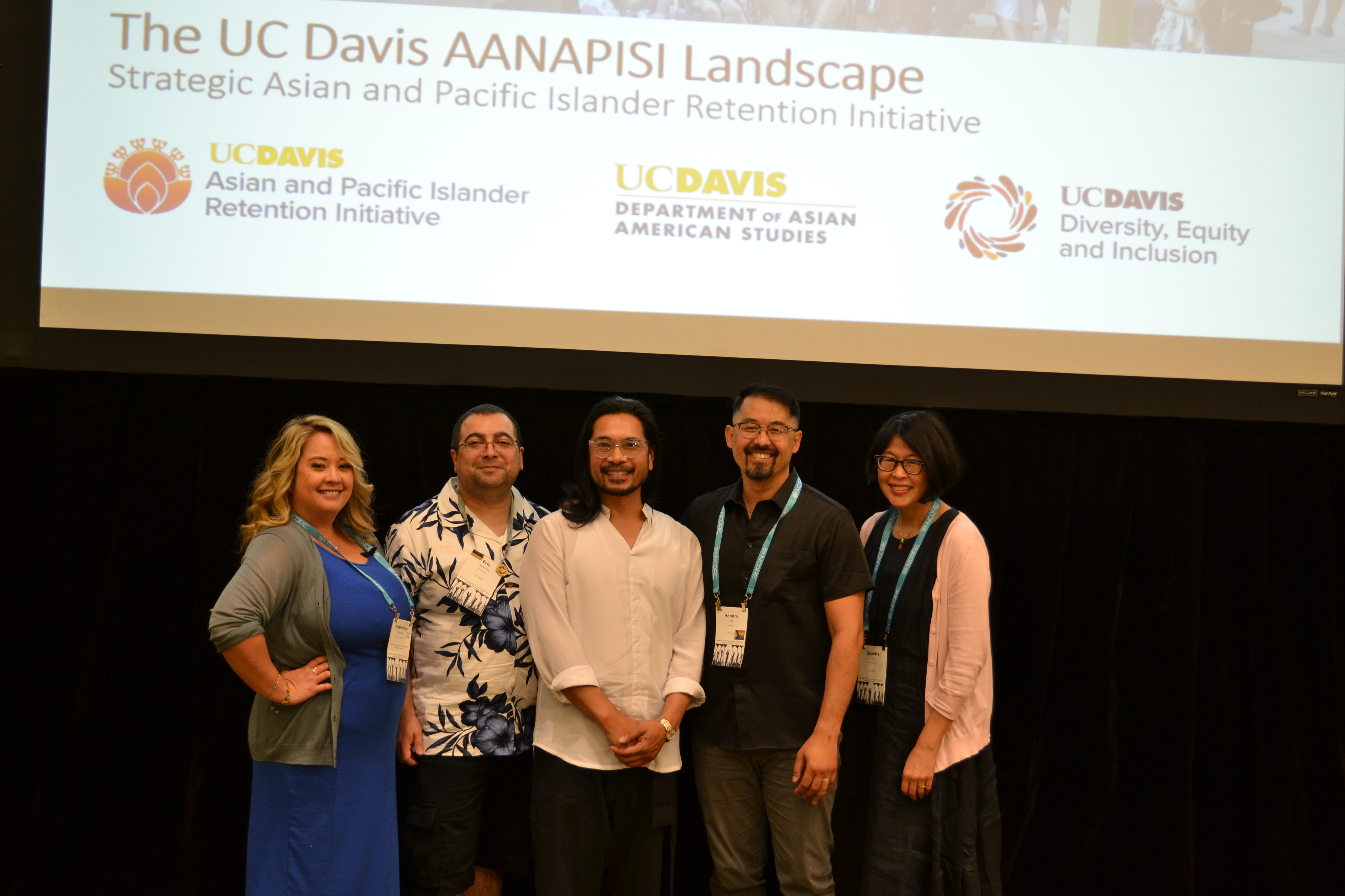 Five people stand in front of a projection screen displaying the UC Davis AANAPISI Landscape initiative.  Text Transcription: "The UC Davis AANAPISI Landscape Strategic Asian and Pacific Islander Retention Initiative" Logos: UC DAVIS Asian and Pacific Islander Retention Initiative UC DAVIS Department of Asian American Studies UC DAVIS Diversity, Equity and Inclusion