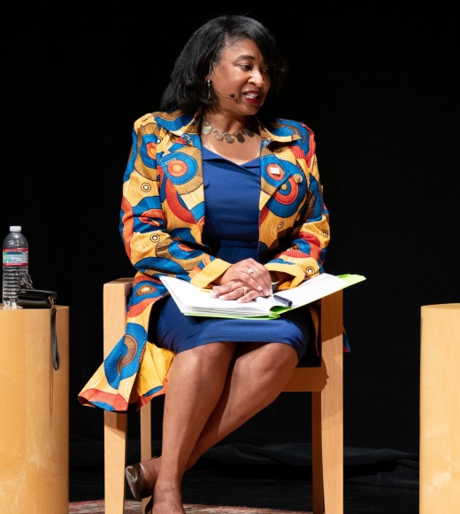 Vice Chancellor Renetta Garrison Tull sits in a chair with an open binder on her lap. She is wearing a blue dress and colorful, long jacket.
