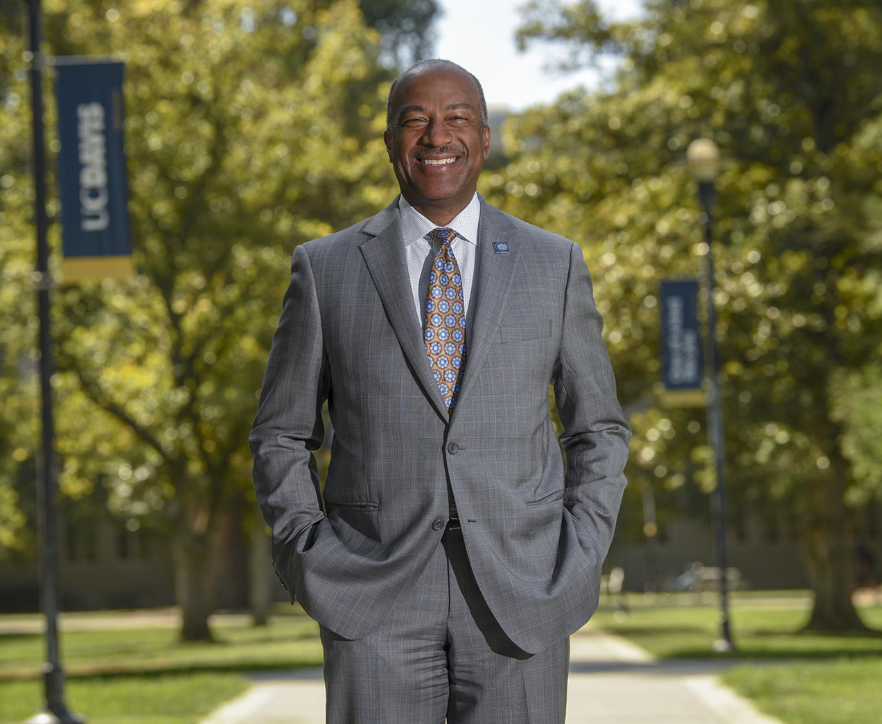Chancellor Gary May smiling and standing on campus with trees and UC. Davis banners behind him