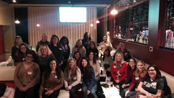 A large group of women, approximately 20, pose together in what appears to be a bar or lounge setting. They're seated and standing in tiers, all smiling at the camera. The room has mood lighting and a projection screen is visible in the background.