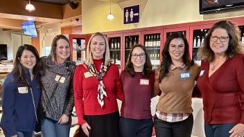 Six women stand together, smiling at the camera. They're in what appears to be a cafe or wine bar, with shelves of wine bottles visible behind them. The women are wearing a variety of colors, with the woman in the center wearing a bright red top. Most are wearing name tags.