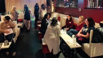 A dimly lit lounge or bar area with women gathered in small groups. Some are seated at tables, others are standing. The space has white couches, red accents, and decorative string lights. Women are engaged in conversation and appear to be networking.