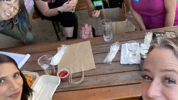 A group of nine women is gathered around a wooden picnic table outdoors, smiling at the camera. There are food containers, drinks, and bags on the table. Some women are seated while others are standing behind them, all appearing to enjoy their time together.