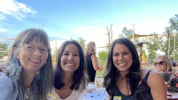 Three women smile at the camera in an outdoor setting. The woman on the left has glasses and blue-tinted hair. The two women on the right have dark hair. Behind them, there's a sunny outdoor area with trees and what looks like playground equipment. Name tags are visible but not legible.