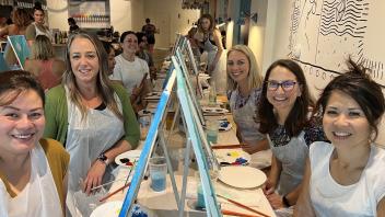  Five women sit at a table with painting supplies, smiling at the camera. They're participating in a painting class or event. The setting appears to be a trendy art studio with bicycles hanging from the ceiling and a decorative wall mural visible in the background.