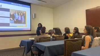 A conference room setting with a presenter standing at the front. The presenter, a woman in a navy top, is gesturing towards a projected slide that reads "Womxn In Tech @ UC Davis". Several attendees are visible from behind, seated at tables with blue tablecloths. Flowers decorate the tables.