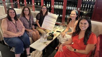 Five women sitting around a small table, smiling at the camera. They are engaged in a planning session, with a large notepad displaying their goals and notes placed on the table in front of them. The background shows shelves filled with wine bottles.