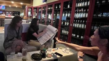  Four women sit at a table in a dimly lit bar or restaurant. Two are looking at a large document while another gestures animatedly. Appetizers and drinks are on the table, and wine bottles are visible in the background.