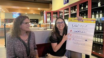 Two women sitting in a wine bar. The woman on the right, wearing glasses, is holding a large notepad with the word 'Celebrate' written at the top, followed by a list that includes 'SWADAAC', 'Events', 'Outreach', and 'Engagement'. She is pointing to the notepad and smiling, while the woman on the left looks on with a smile.