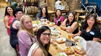 A group of eight women are seated around a wooden table enjoying a meal together at a brewery or casual dining spot. The setting includes barrels and industrial equipment in the background. They are smiling, with some wearing name tags. Various drinks and plates of food like nachos and dips are spread across the table, creating a relaxed and friendly atmosphere.