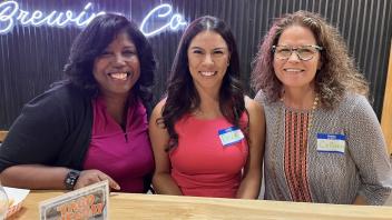 Three women are seated together at a wooden table in a brewery with a neon sign in the background. They are smiling, with name tags reading "Sonja," "Erica," and "Colleen." The woman on the left is wearing a pink top, the middle woman is in a sleeveless coral dress, and the woman on the right is wearing a patterned top with glasses. 