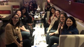 A group of about 15 women seated around tables in a lounge-like setting. They are smiling at the camera, with papers and drinks on the tables. The group is diverse, and some are wearing name tags. The setting suggests a casual yet professional gathering.