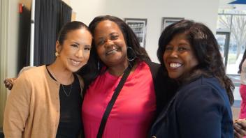 Three women standing together and smiling at the camera. From left to right: a woman in a tan cardigan, a woman in a bright pink top, and a woman in a navy blazer. They appear to be at a professional event, with a backdrop of office or conference room visibl