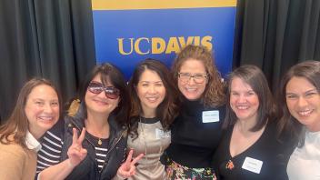 A group of six women is smiling and posing together in front of a UC Davis banner. Two women in the group are making peace signs with their fingers. All are dressed in semi-formal attire and appear to be at an event or gathering  Six women posing together in front of a UC Davis branded backdrop. They're all smiling, and two are making peace signs with their hands. The women appear diverse in age and ethnicity. They're dressed in casual to business casual attire, and some are wearing name tags.
