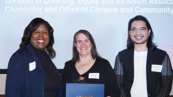 Three smiling people stand together at an event. On the left, a woman with curly hair wears a navy blazer and a name tag. In the middle, a woman with shoulder-length hair holds a blue folder with the UC Davis logo and wears a black top and a name tag. On the right, a person with long dark hair and glasses wears a white shirt and patterned cardigan. Above them, a banner partially reads "Division of Diversity, Equity and Inclusion Associate Chancellor and Office of Campus and Community".