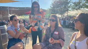 Four women are gathered at an outdoor event with string lights overhead. They're wearing casual clothes and sunglasses. One woman is standing, holding a drink, while the others are seated. The woman wearing a name tag that says "Colleen" is holding a glass. In the background, a crowd of people can be seen enjoying the event.