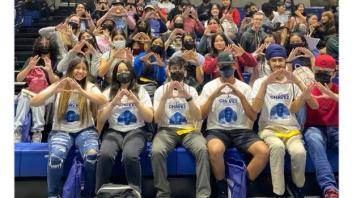 A crowd gathers in an auditorium wearing their CCYLC shirts. In the front row, five students hold up their fingers to make the shape of a heart.