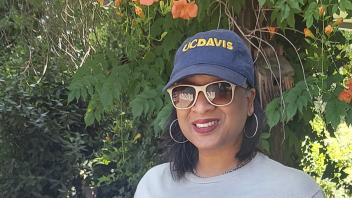 Renetta Tull stands in front of an autumn landscape, wearing sunglasses and decked out in a blue hat and gray t-shirt with the UC Davis Diversity, Equity and Inclusion, logo. 