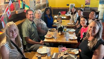 A group of ten people are seated around a long table in a restaurant. Various international flags hang on the wall behind them. The group appears diverse in age, gender, and ethnicity. They are all smiling at the camera, with plates of food and drinks in front of them. The setting suggests a casual, friendly gathering.