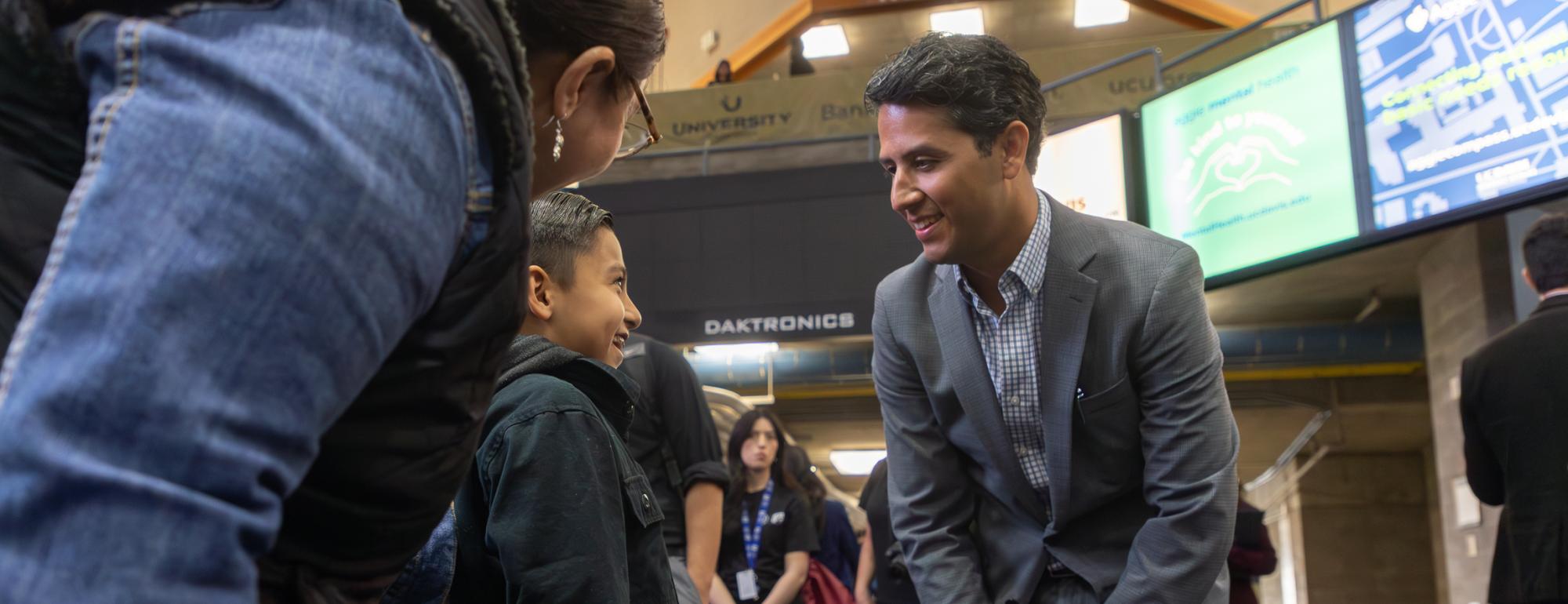 A man in a gray suit smiles at a young boy in a dark jacket in an indoor area with digital signage.