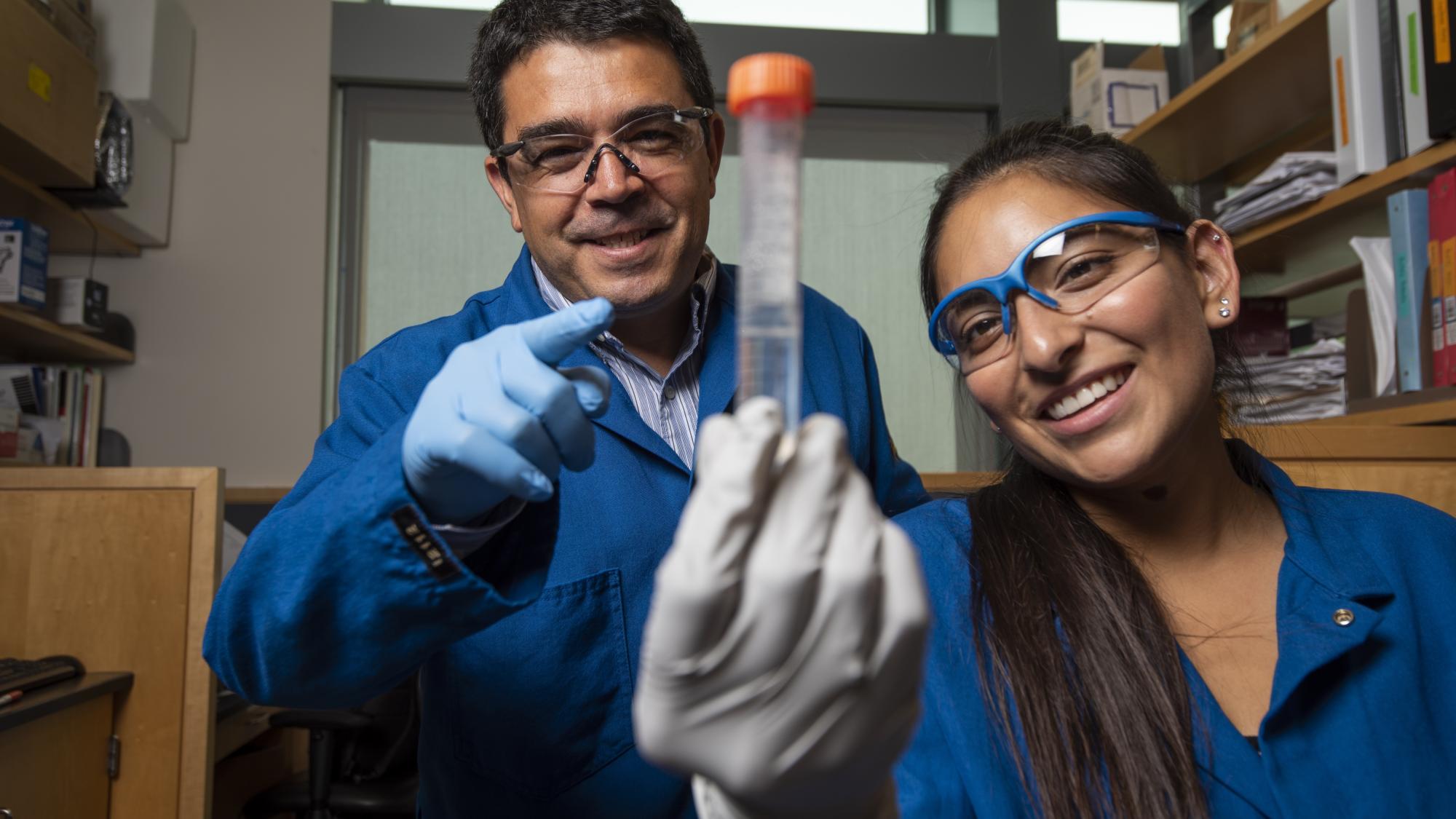 Two lab technicians in blue lab coats and safety goggles examining a test tube.