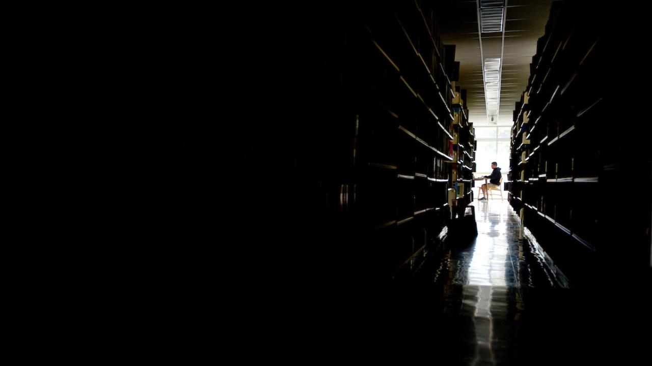 The library stacks in shadows frame a single person studying in front of a brightly lit window.