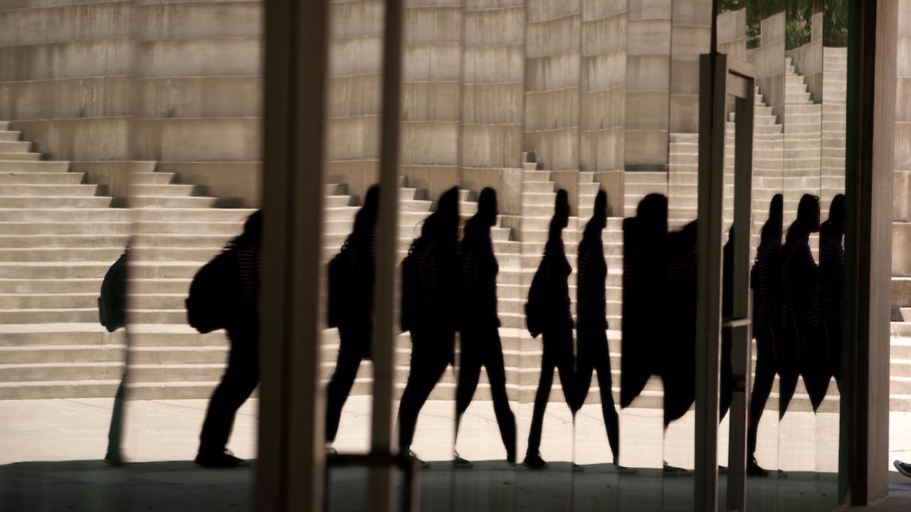 Reflections of people walking, distorted in a mirrored building surface with a background of concrete steps and walls.