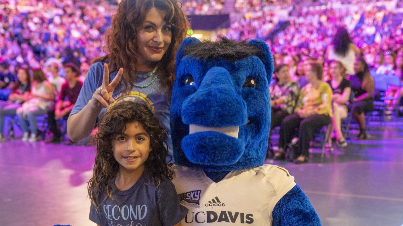 Woman and child posing with a blue furry mascot at a crowded indoor event. Transcribed Text on child's t-shirt says SECOND GRADE ROCKS. Transcribed text on mascot's jersey says UC DAVIS 19