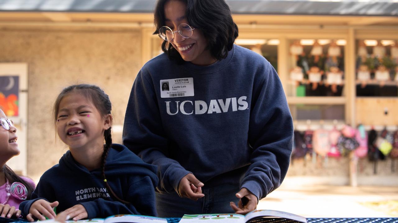 Person in UC Davis sweatshirt smiling at children at a table outdoors.