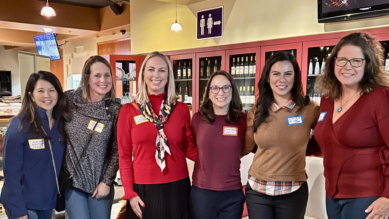 Six women stand together, smiling at the camera. They're in what appears to be a cafe or wine bar, with shelves of wine bottles visible behind them. The women are wearing a variety of colors, with the woman in the center wearing a bright red top. Most are wearing name tags.