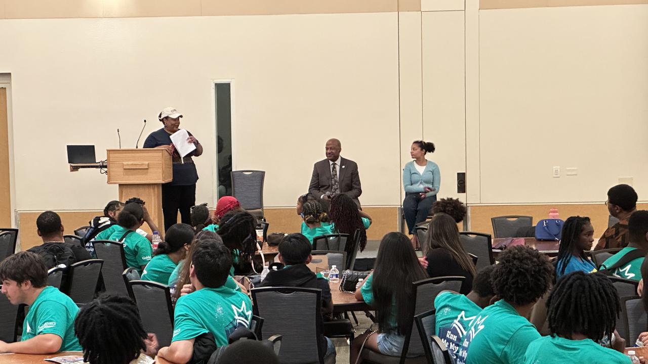 Chancellor May sits on stage in front of crowd of middle school and high school students seated at round tables wearing bright green tshirts. Ebony Lewis is at podium talking to crowd.