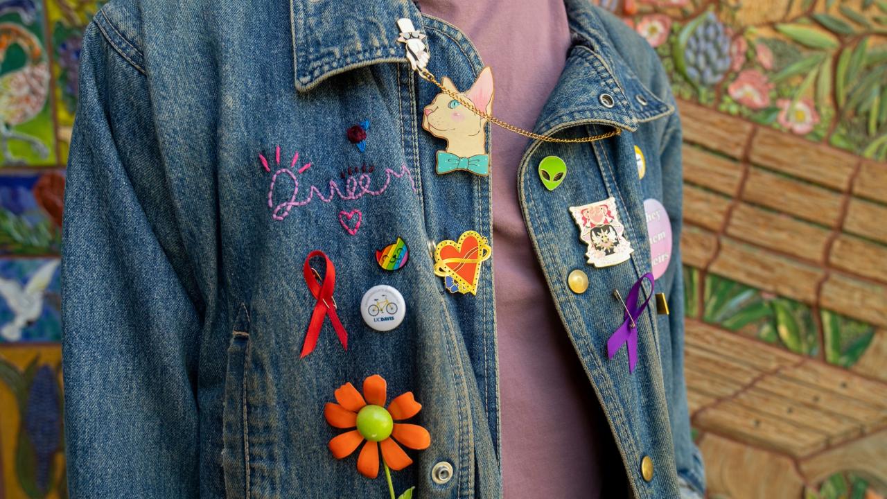 A person cropped from neck to waist wearing a jean jacket with stitching that reads "Queer," buttons that represent UC Davis, a red ribbon, a purple ribbon, a heart, a cat, a daisy, and a button that says "they, them, theirs"  