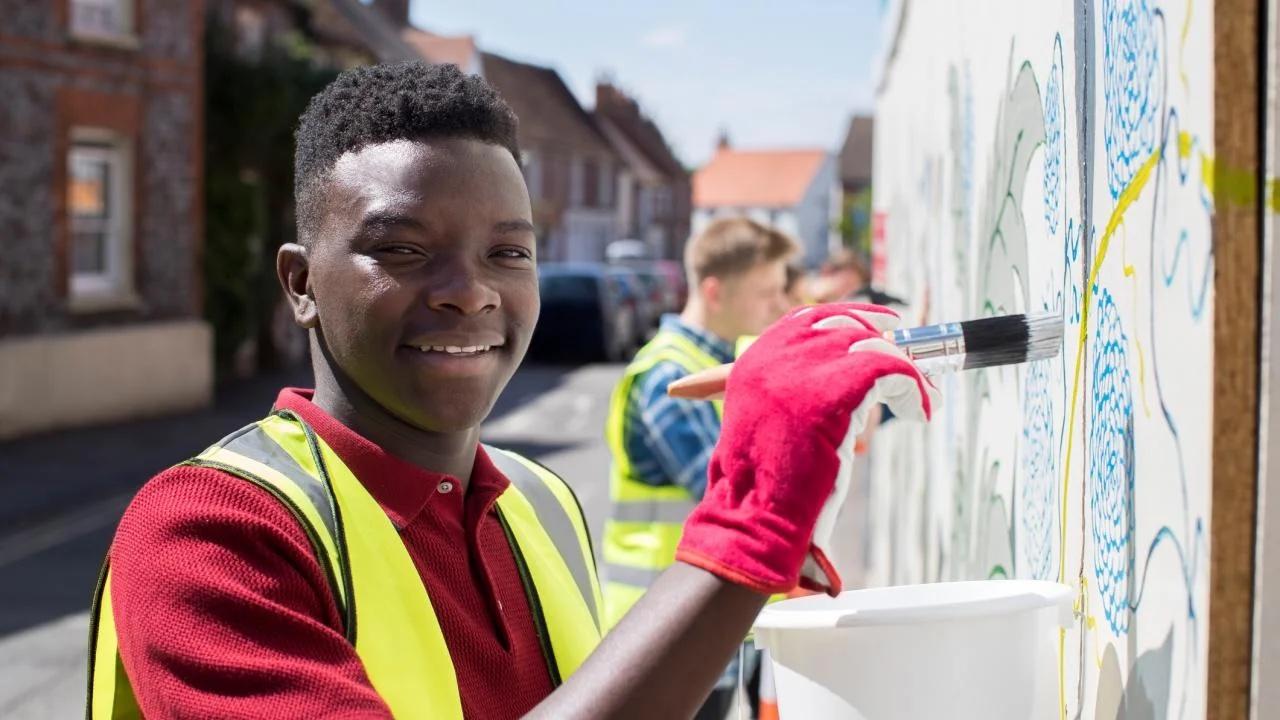 African American teenager wearing a red polo shirt, bright yellow construction vest and red gloves is painting a mural.