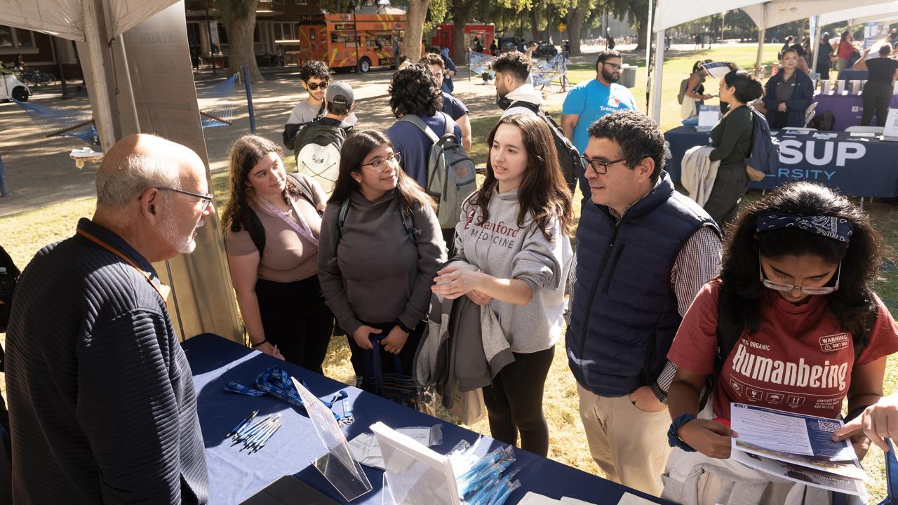 Three female students talk with two male professors at tabling event on UC Davis quad. Another female student is reading a brochure.