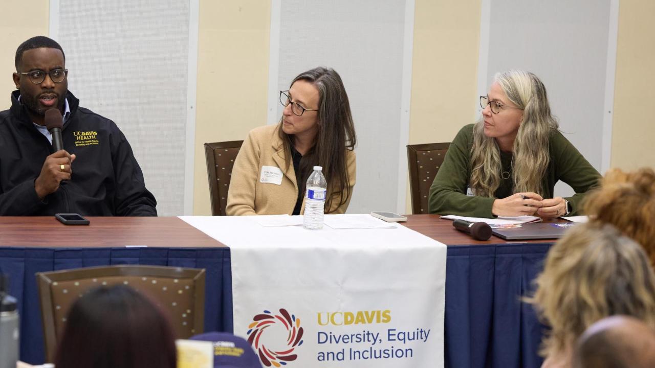 African American man sits at table and speaks into microphone to crowd seated at round tables. Two women, who are seated next to him, look at him as he speaks.