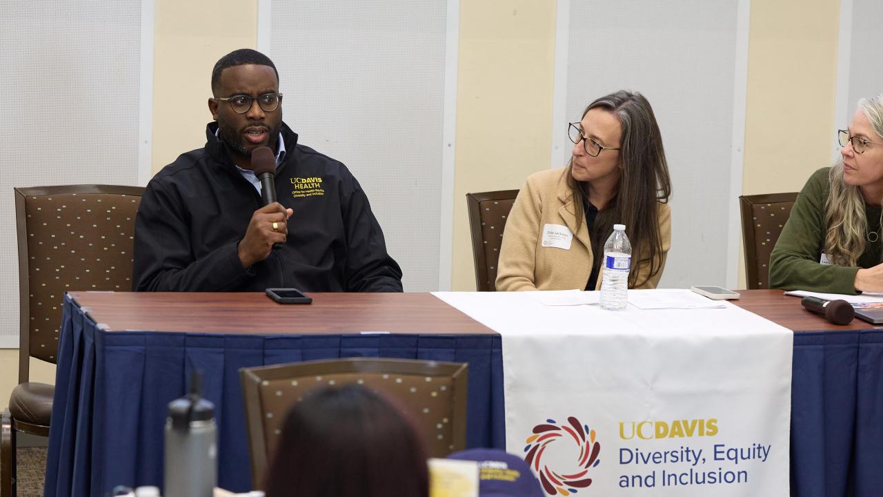 African American man sits at table and speaks into microphone to crowd seated at round tables. Two women, who are seated next to him, look at him as he speaks.