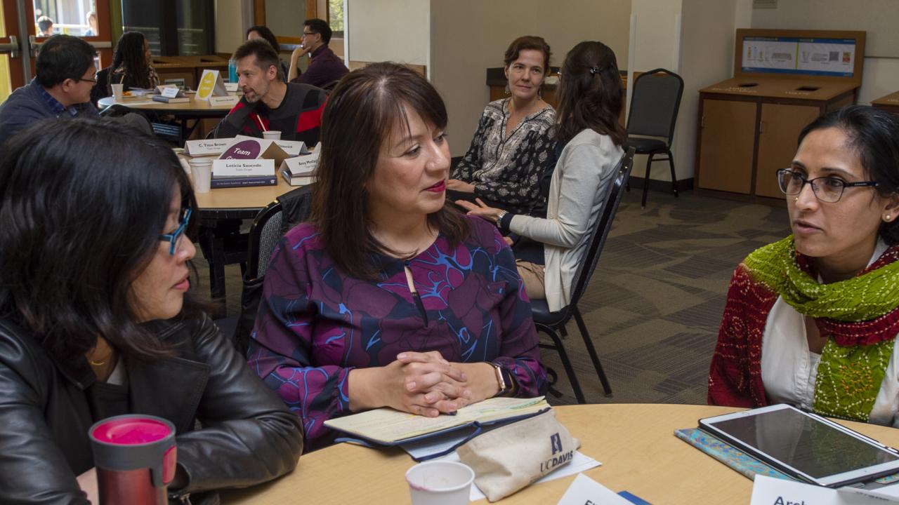 Three women sit around a table engaged in conversation. In the background, other women and men are seated at round tables engaged in coversation.