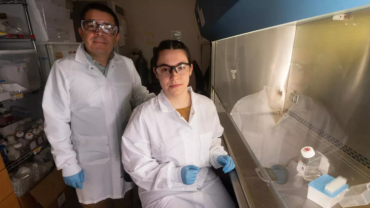 Fifth-year student Jasmine Diaz (seated right) and Professor Luis Carvajal-Carmona (standing left) pictured in lab wearing white lab coats, gloves and protective goggles.