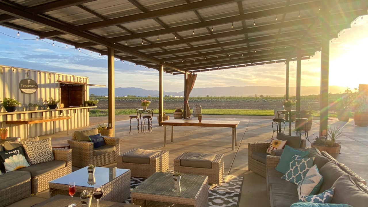 Couches and tables under a pergola overlooking a vineyard at sunset.