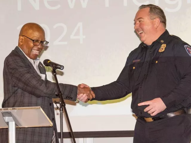 Two men shake hands on stage. One man is wearing a suit, the other is wearing a police uniform.