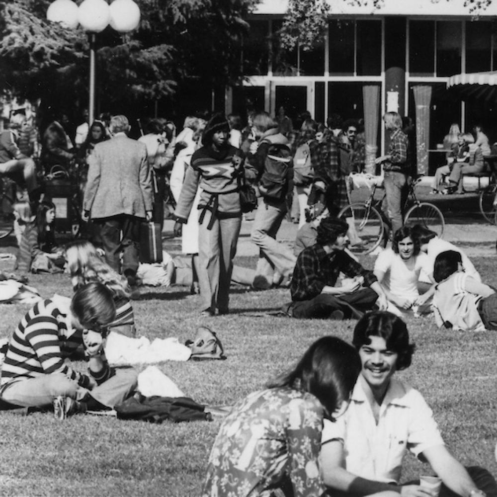 Black and white photo of students gathered on a campus lawn, with buildings and trees in the background.