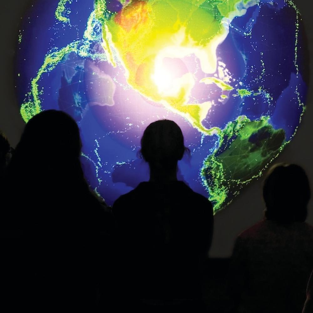 Silhouetted figures in front of a brightly colored digital image of Earth on a screen.