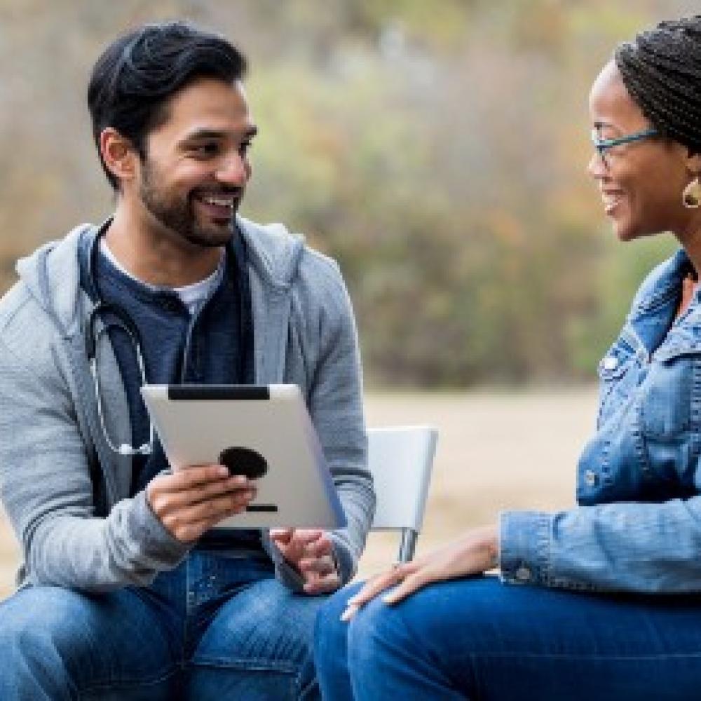 A man with a stethoscope and a tablet talks to a woman in a denim jacket outdoors.