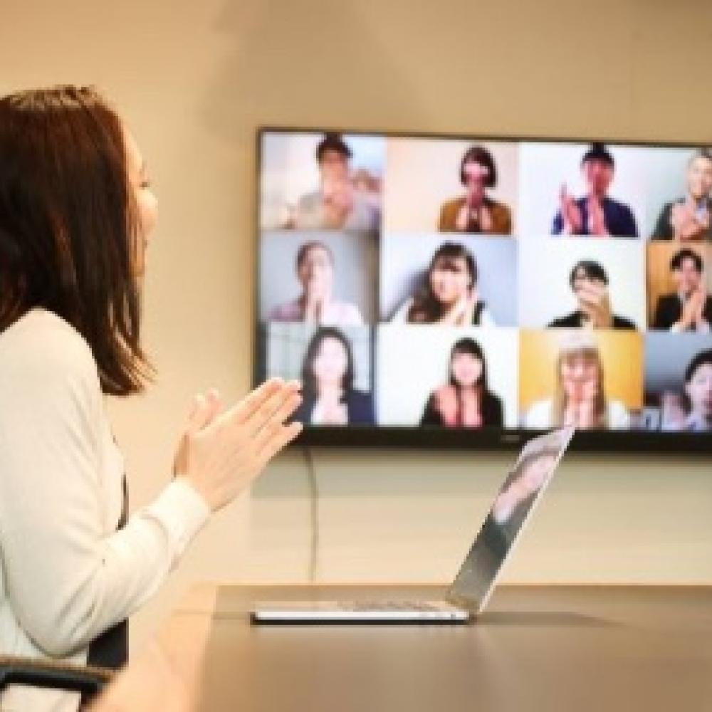 A woman participates in a video call, facing her laptop and a large screen displaying multiple call participants.