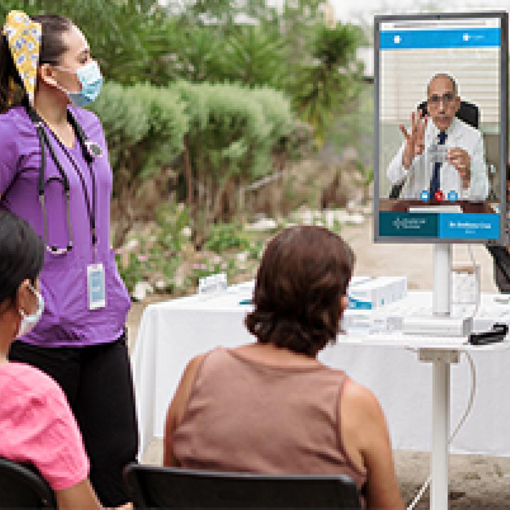 A healthcare worker stands beside two people watching a telemedicine consultation on a screen outdoors.