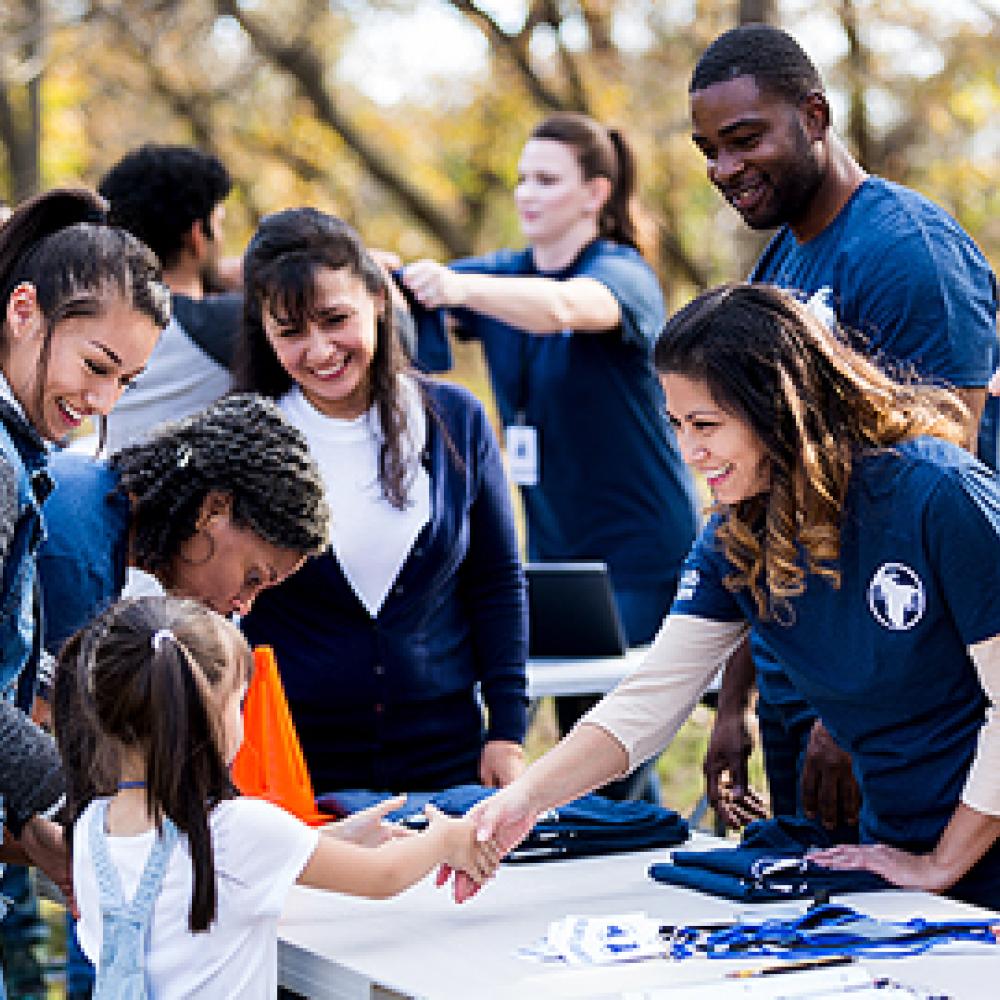 A group of people in blue shirts interacting with a child across a table in an outdoor setting.