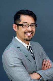 Asian man with glasses and a grey sports coat, folds arms and looks over his shoulder at the camera.