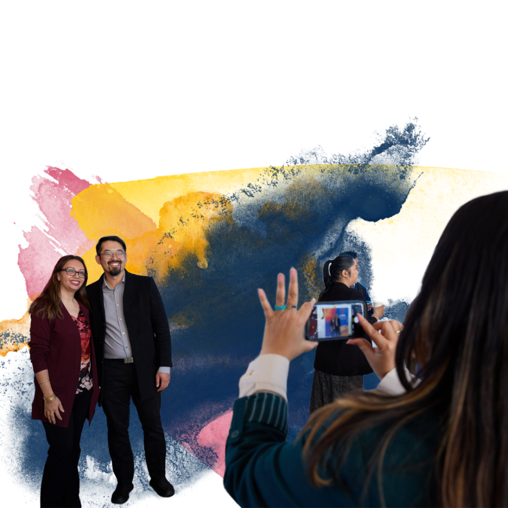 Two people pose for a picture in front of a colorful watercolor background, with another person taking their picture