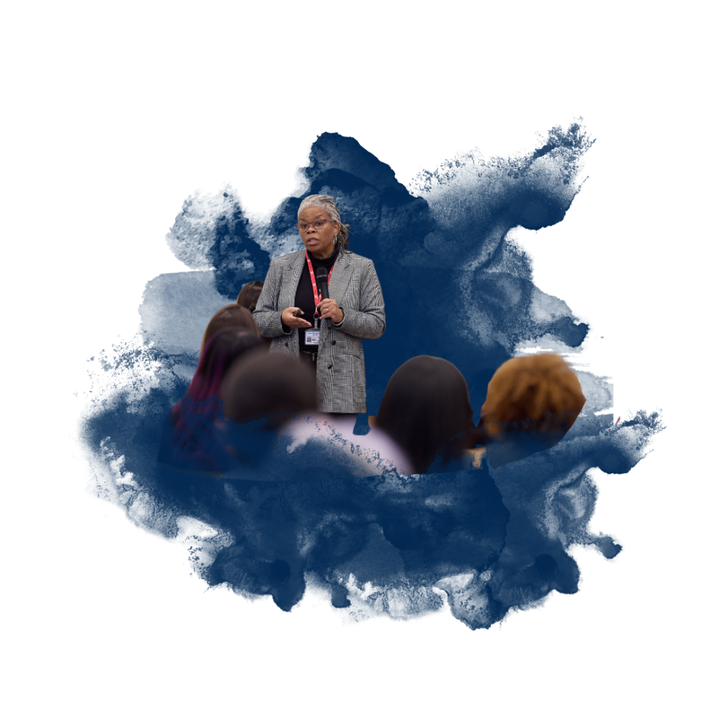 A woman in a gray blazer speaks to an audience against a blue watercolor splash background.